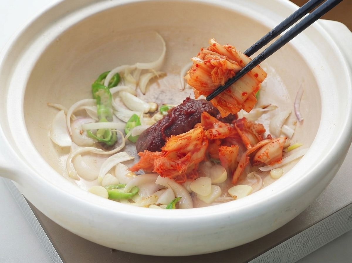 Pasta de chile coreano y kimchi siendo añadidos a la olla de aromáticos usando palillos.