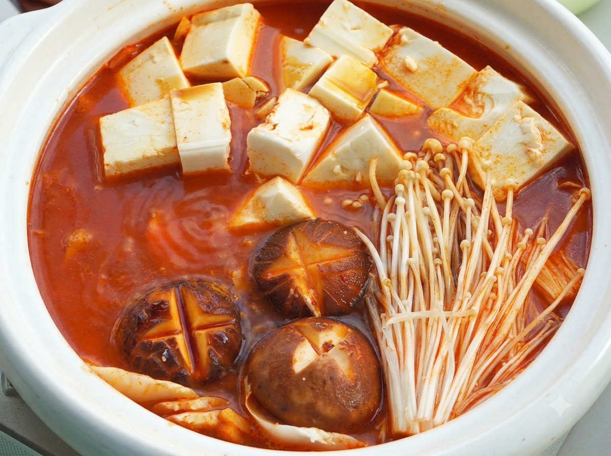 Una olla caliente de cerámica blanca llena de base de sopa roja hirviendo, cubos de tofu suave, champiñones shiitake con cortes en cruz y setas enoki bajo una tapa.