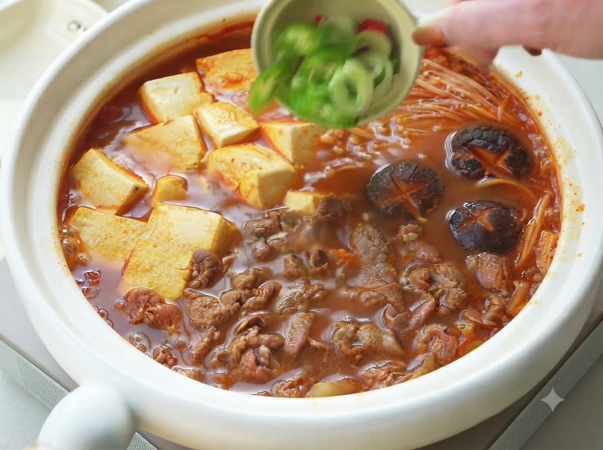 Cebolletas y chiles cortados vertiéndose desde un tazón pequeño como guarnición sobre el estofado coreano de carne y tofu ya cocido.