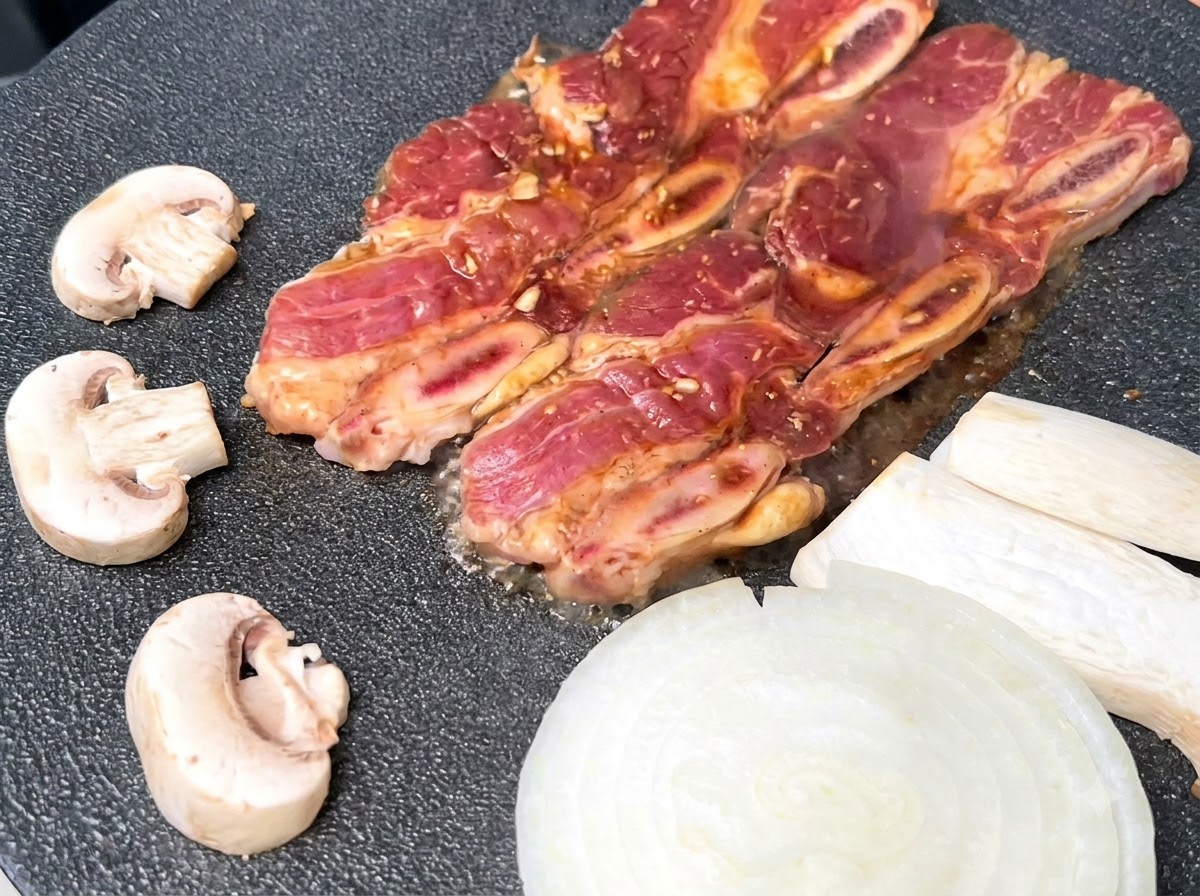 Costillas cortas de res marinadas chisporroteando en una sartén grill negra caliente junto a cebollas blancas en rodajas y champiñones.
