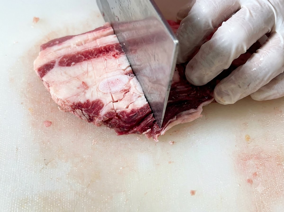 Costillas cortas de res crudas siendo cortadas en piezas rectangulares uniformes sobre una tabla de picar usando un cuchillo grande.