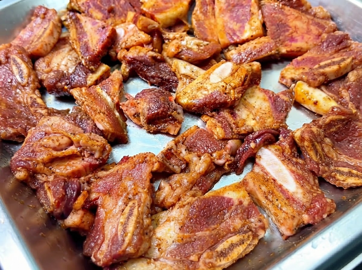 Costillas cortas de res marinadas dispuestas en una bandeja de metal con cebollas trituradas espolvoreadas encima.