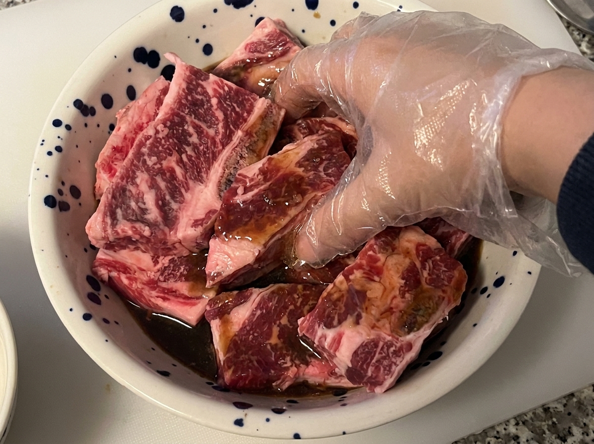 Una mano con guante mezclando y masajeando costillas de res crudas con adobo oscuro en un tazón.