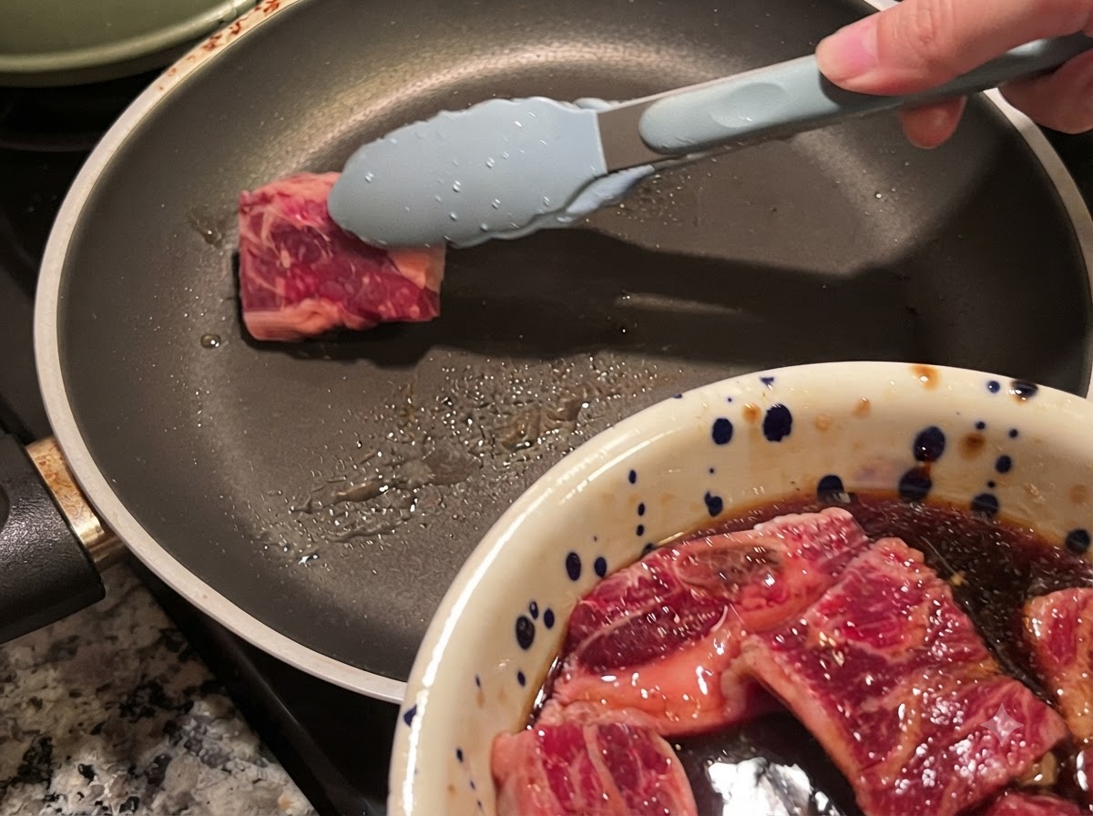 Pinzas colocando una pieza de costilla de res marinada en una sartén caliente.