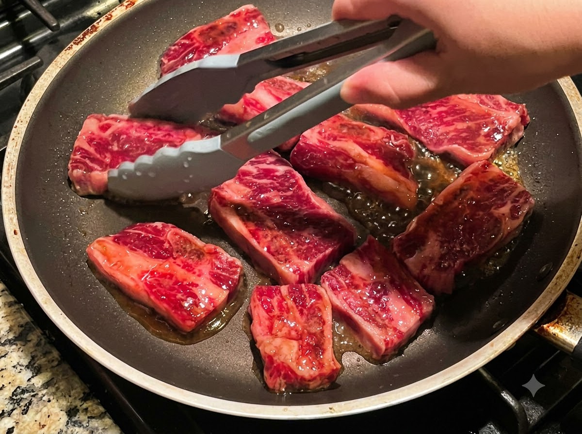Pinzas volteando múltiples piezas de costillas de res friéndose en una sartén.