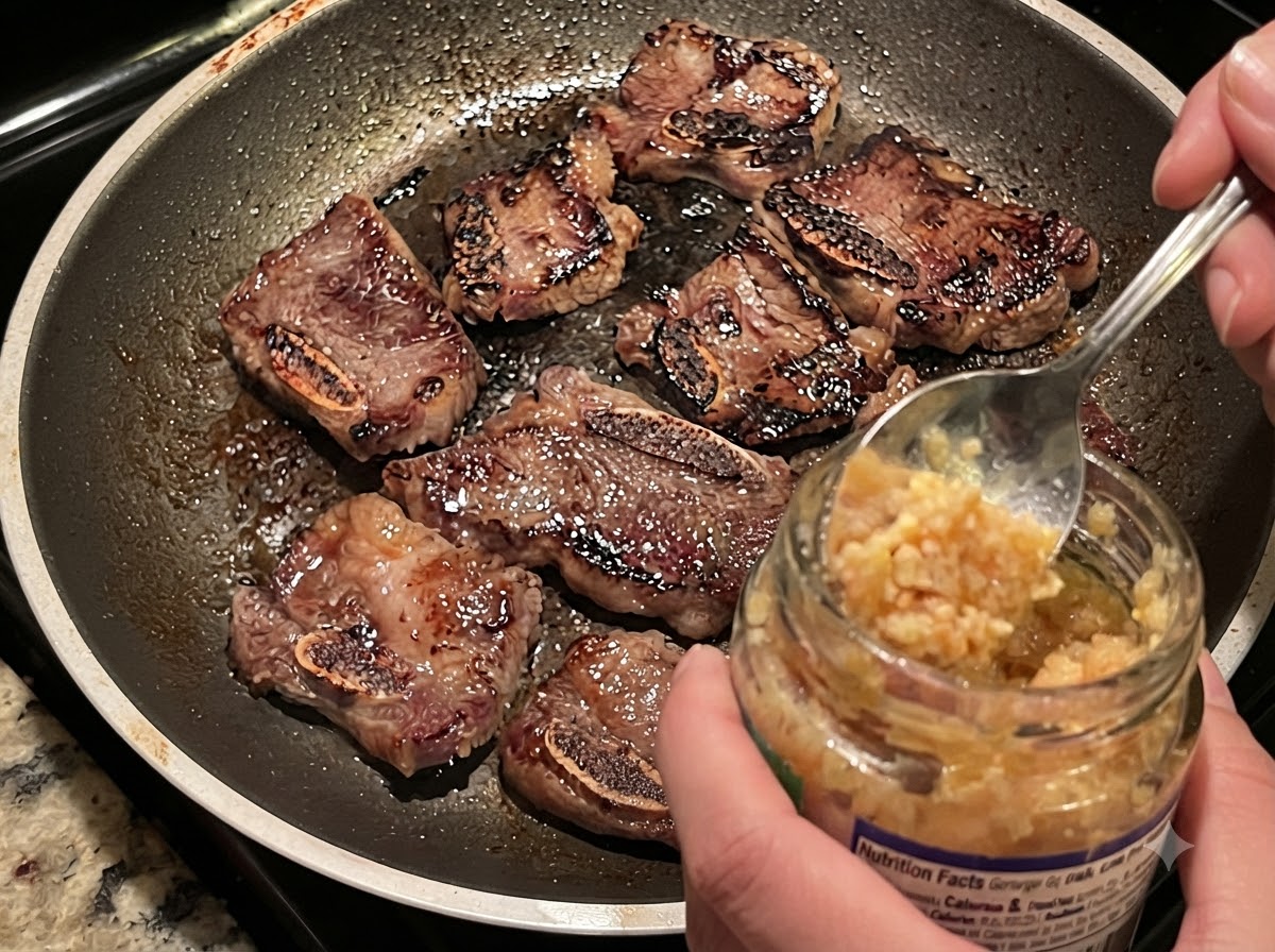 Ajo picado siendo agregado con una cuchara desde un frasco hacia una sartén caliente con costillas de res dorándose y cebollas.