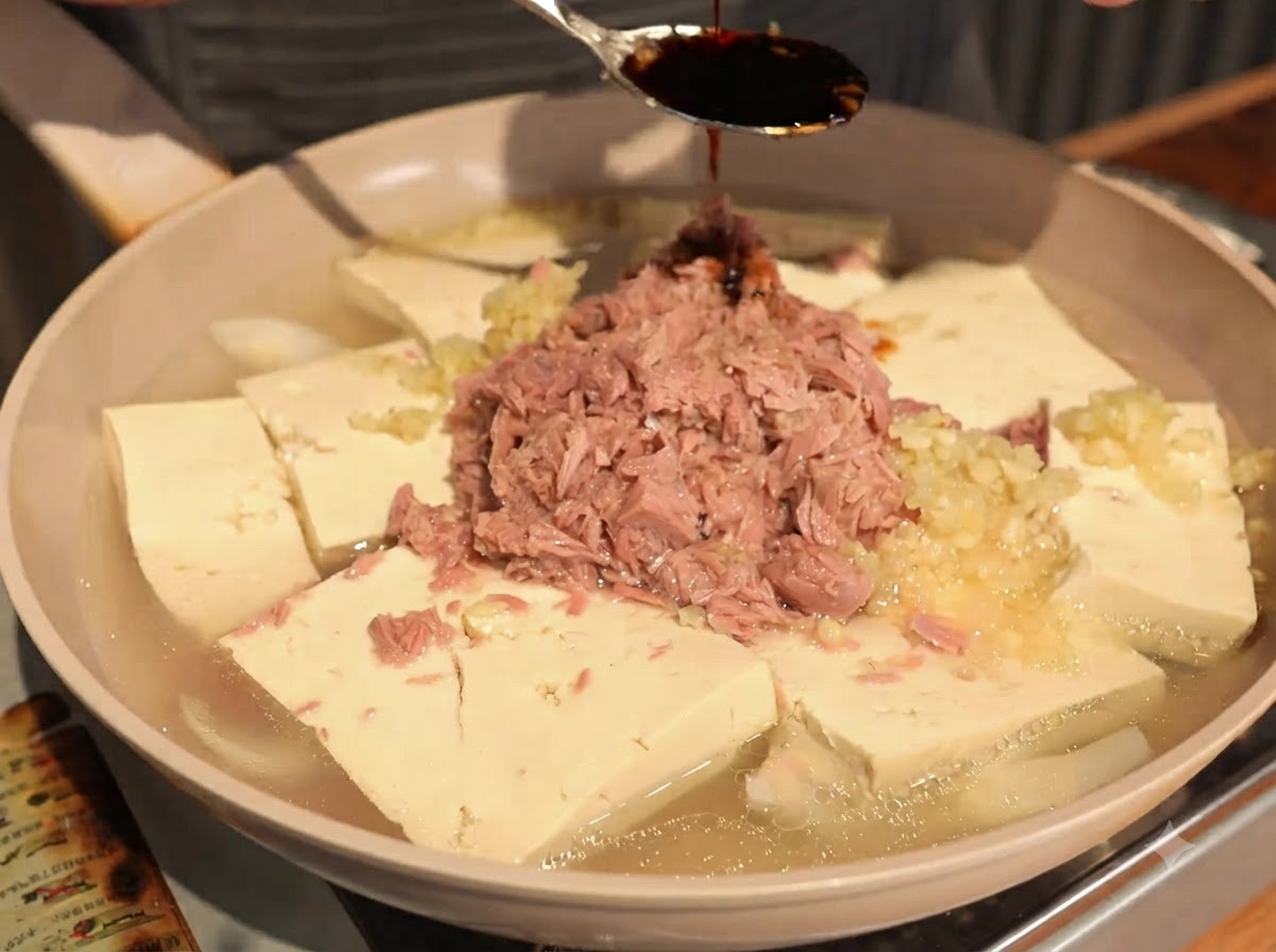 Salsa de soya oscura siendo rociada desde una cuchara sobre el centro del atún en lata y las piezas de tofu.