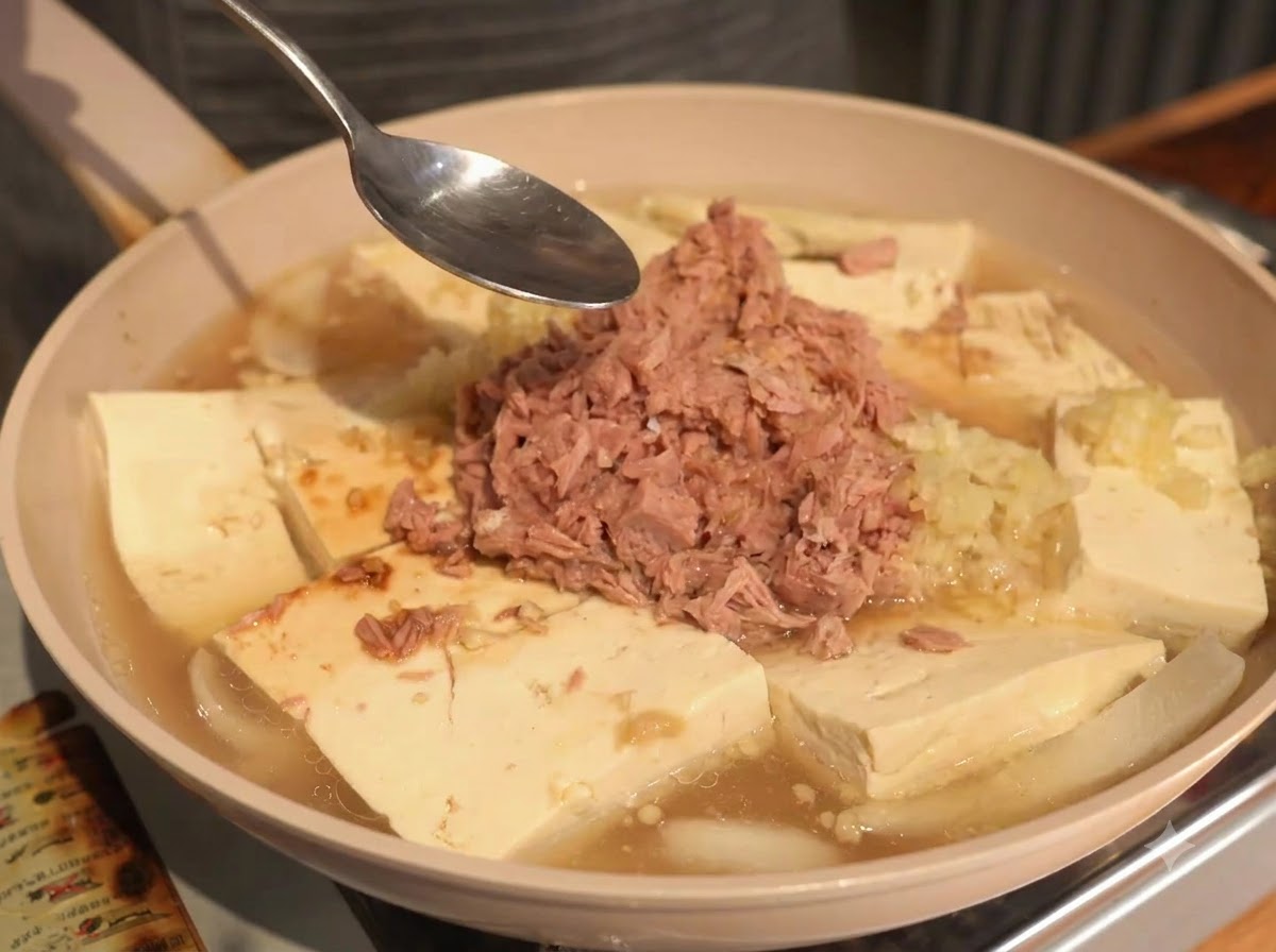 Una cuchara sostenida sobre la sartén que contiene atún, tofu y caldo líquido durante el proceso de condimentación.