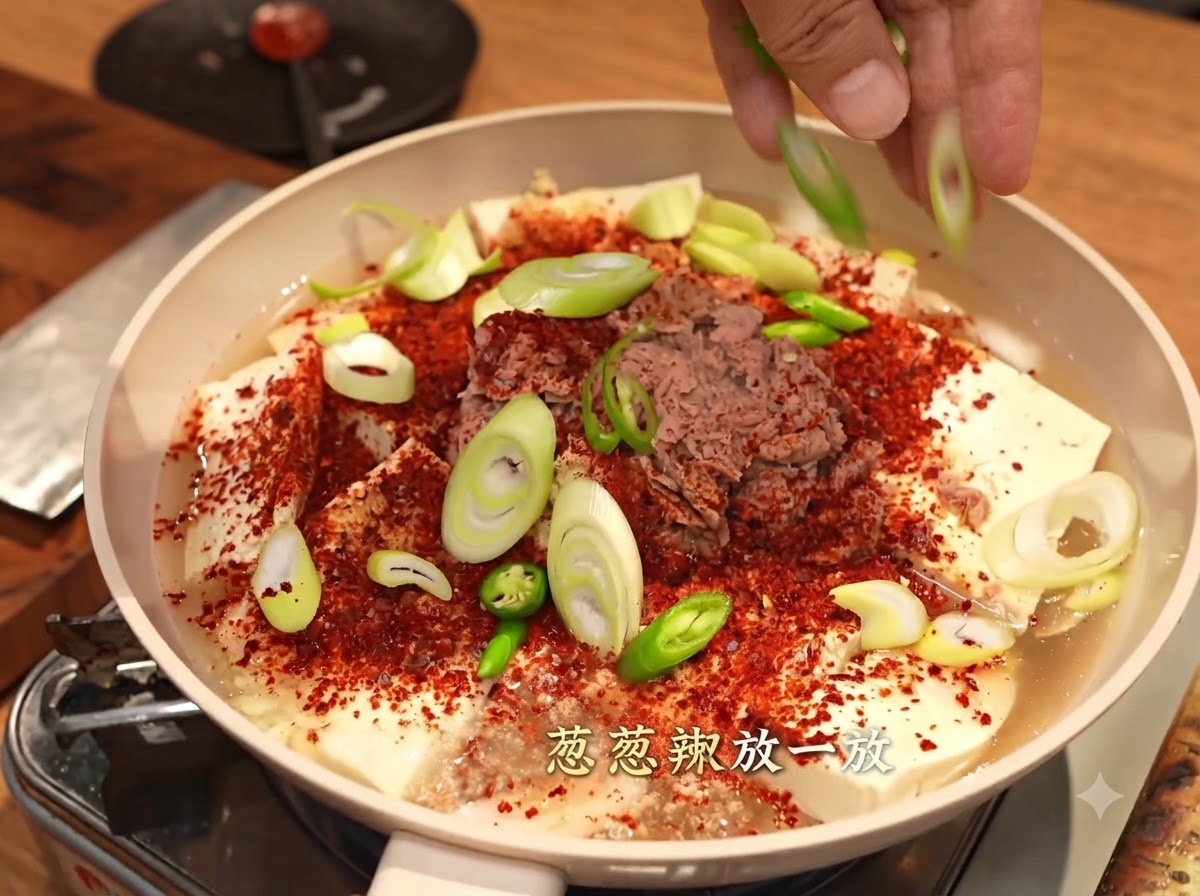 Manos esparciendo cebollines cortados y chiles verdes sobre una sartén cubierta con hojuelas de chile rojo grueso, tofu y atún.