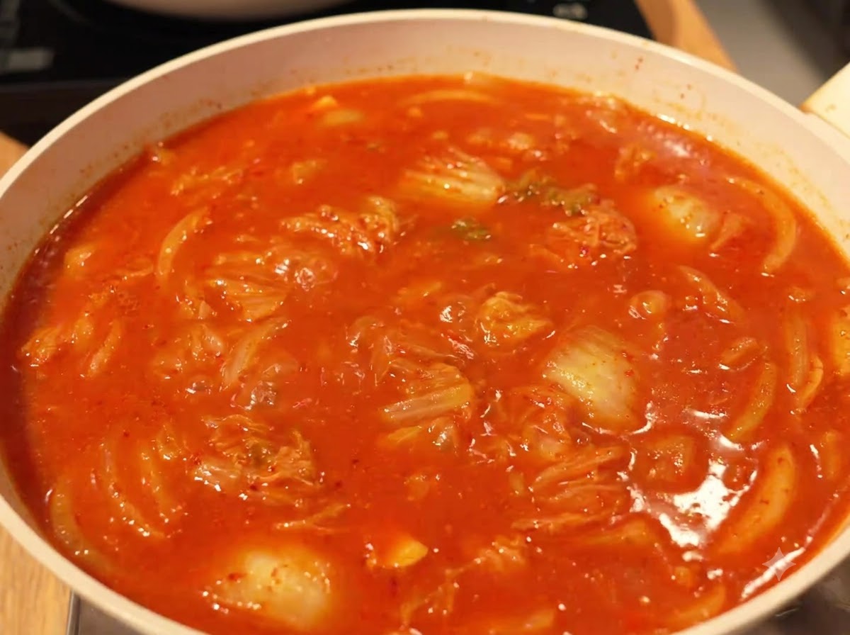 Un caldo rojo rico y burbujeante cocinándose a fuego lento con kimchi e ingredientes en una sartén en la estufa.