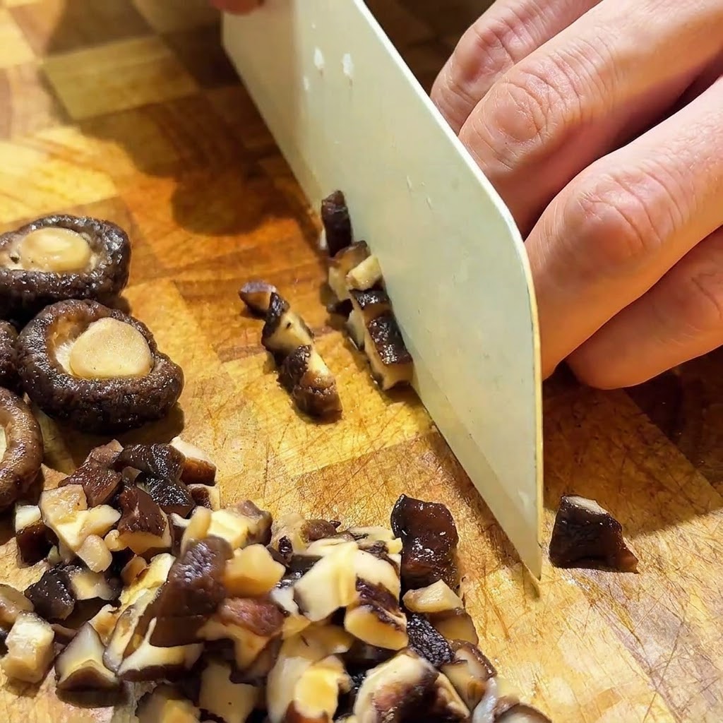 Setas shiitake rehidratadas siendo cortadas en pequeños cubos con un cuchillo blanco sobre una tabla de madera.