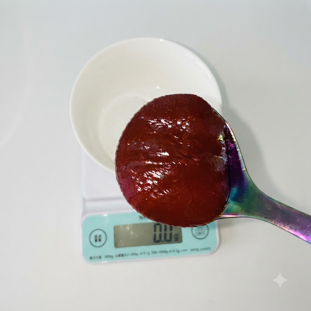 Una cuchara con pasta de chile rojo coreana sostenida sobre un tazón blanco que descansa en una báscula de cocina digital.