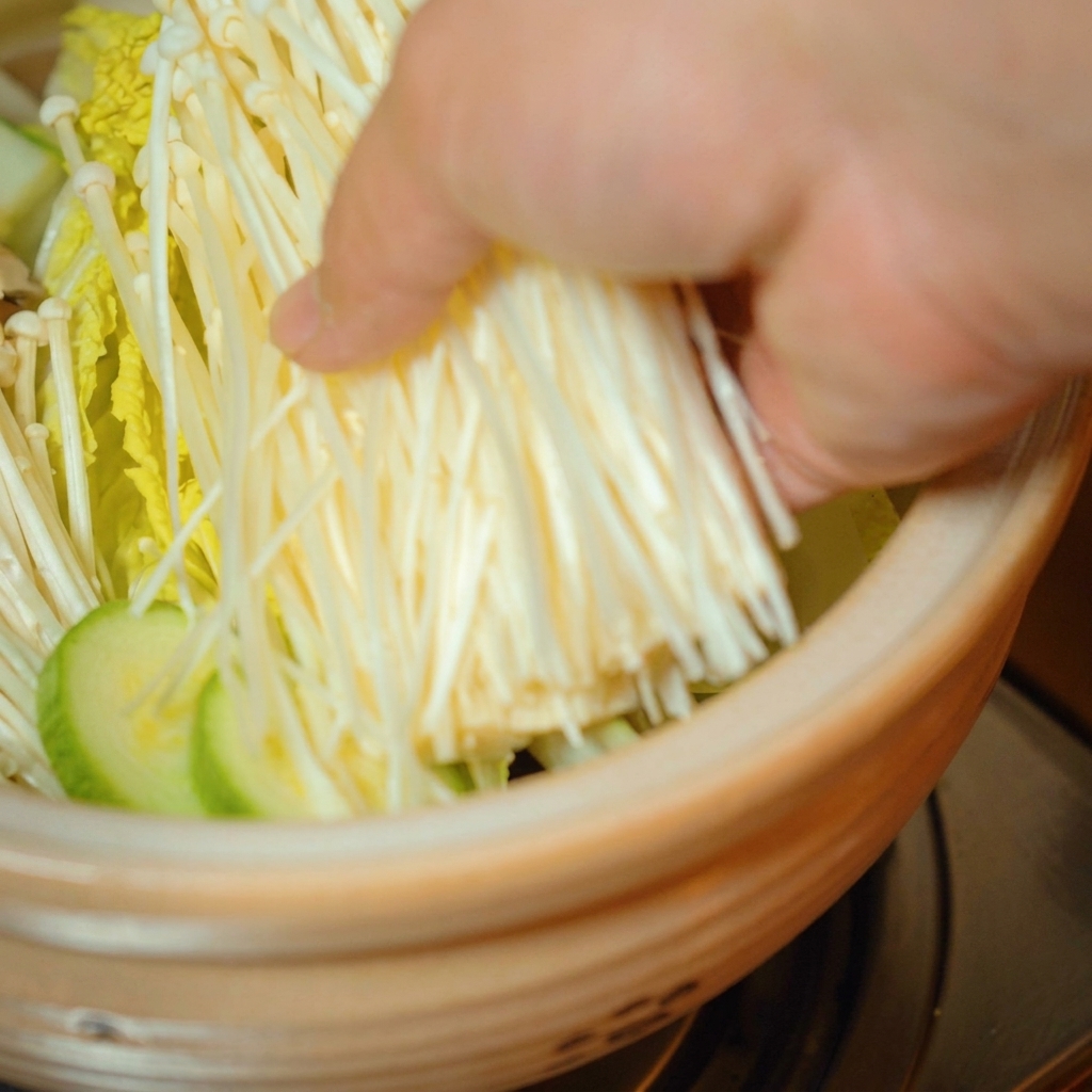 Hongos enoki frescos siendo colocados encima de las verduras en la olla.