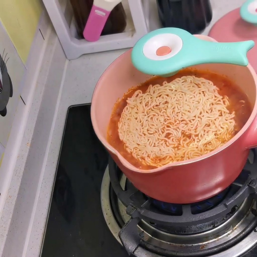 Un paquete de fideos instantáneos siendo arrojado en una olla rosa de caldo rojo picante hirviendo en una estufa.