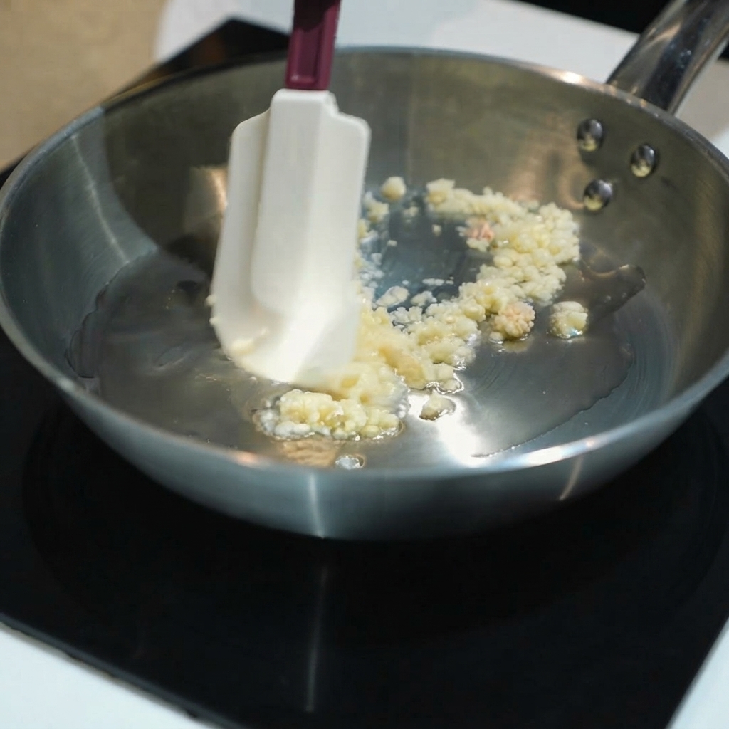 Ajo picado siendo salteado en aceite dentro de una sartén de acero inoxidable usando una espátula de silicona.