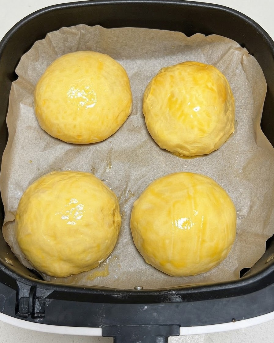Cuatro panecillos de masa de hamburguesa fermentados cubiertos con un baño de huevo brillante dentro de la canasta de la freidora de aire.