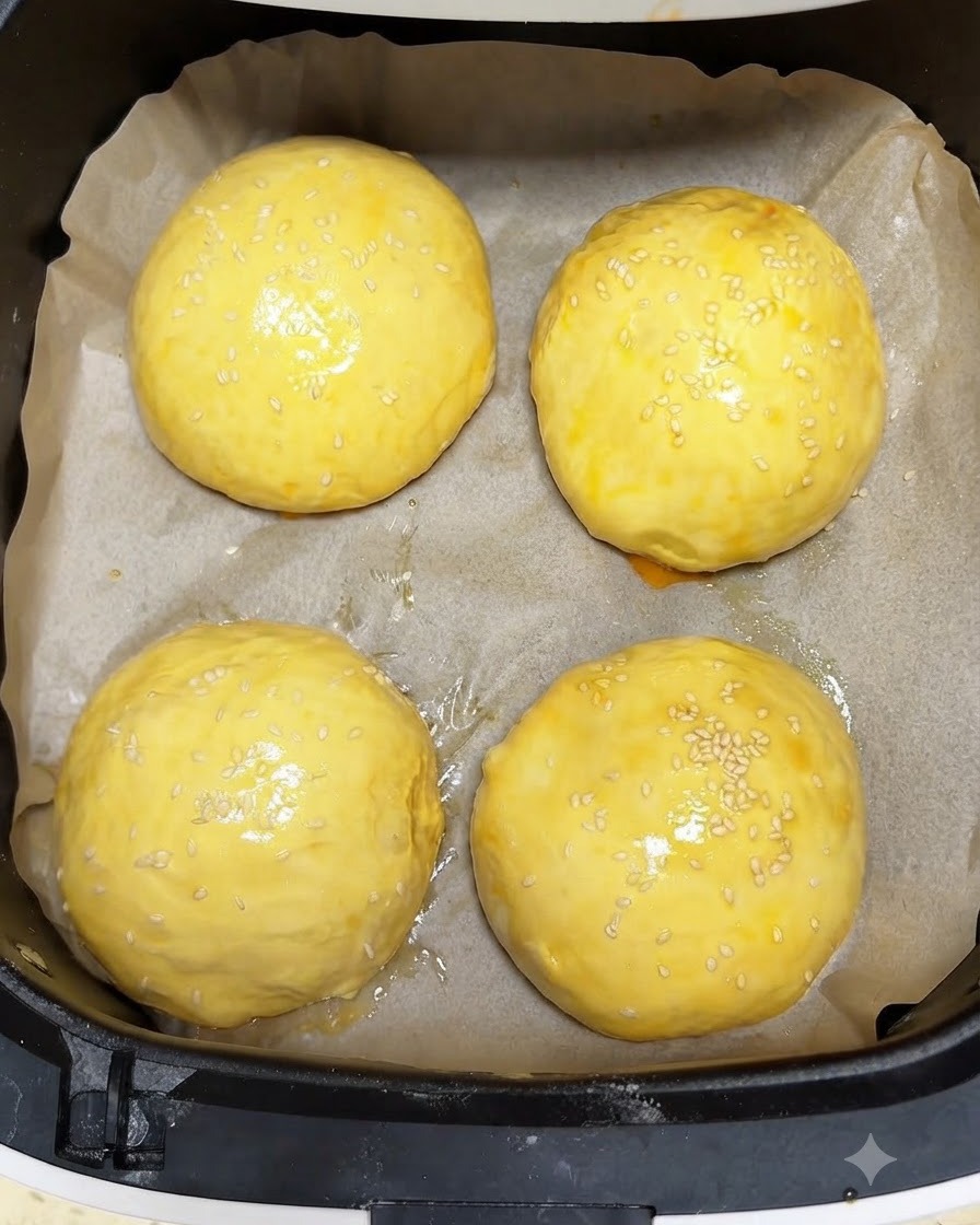 Cuatro panecillos de masa dorados con baño de huevo y cubiertos con semillas de sésamo dentro de una canasta de freidora de aire, listos para hornear.