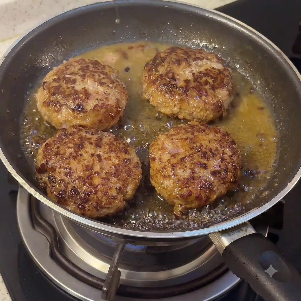 Un bloque de mantequilla y componentes de salsa derritiéndose en la sartén con las hamburguesas chisporroteantes.