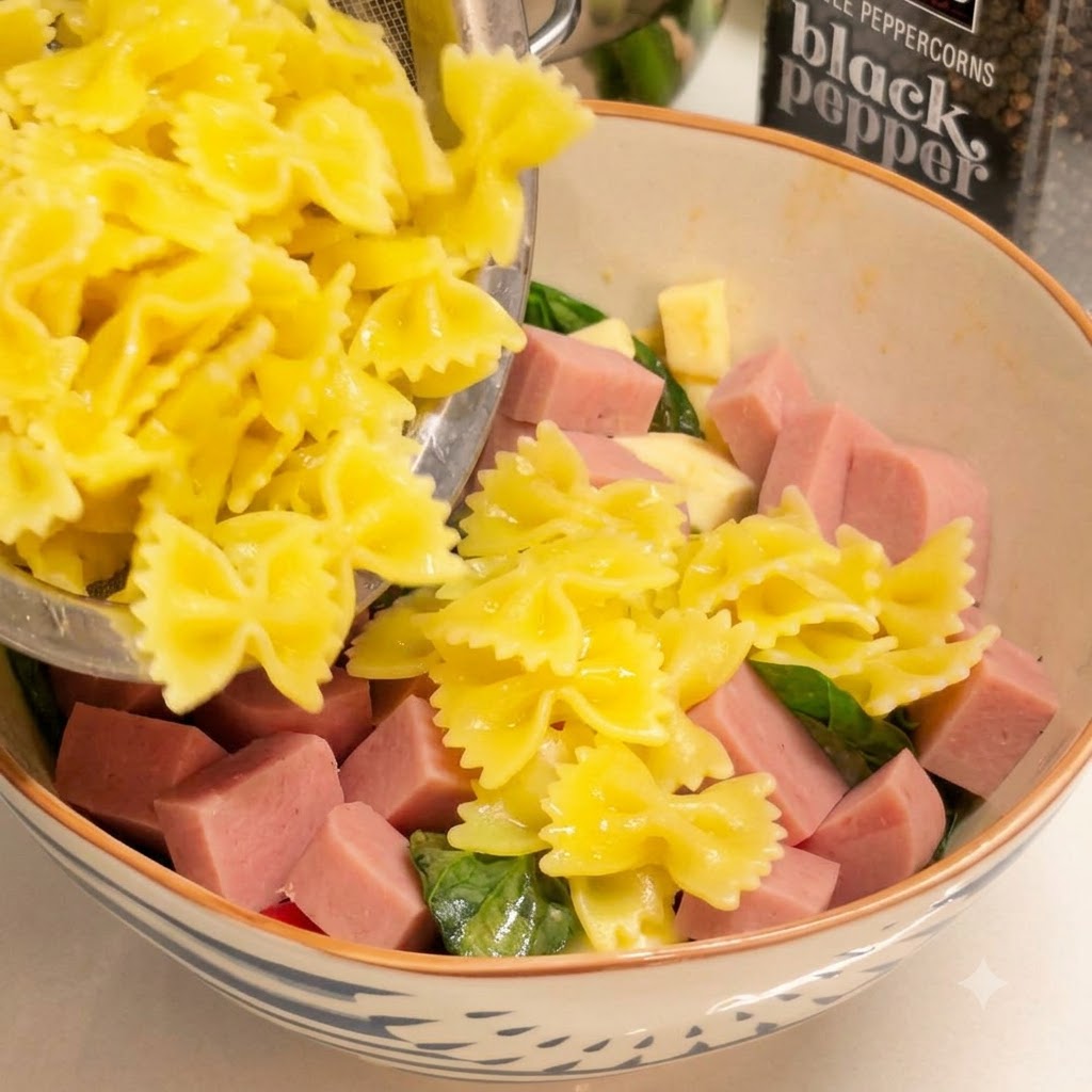 Pasta farfalle amarilla cocida siendo escurrida y transferida a un bol con jamón en cubos y verduras.