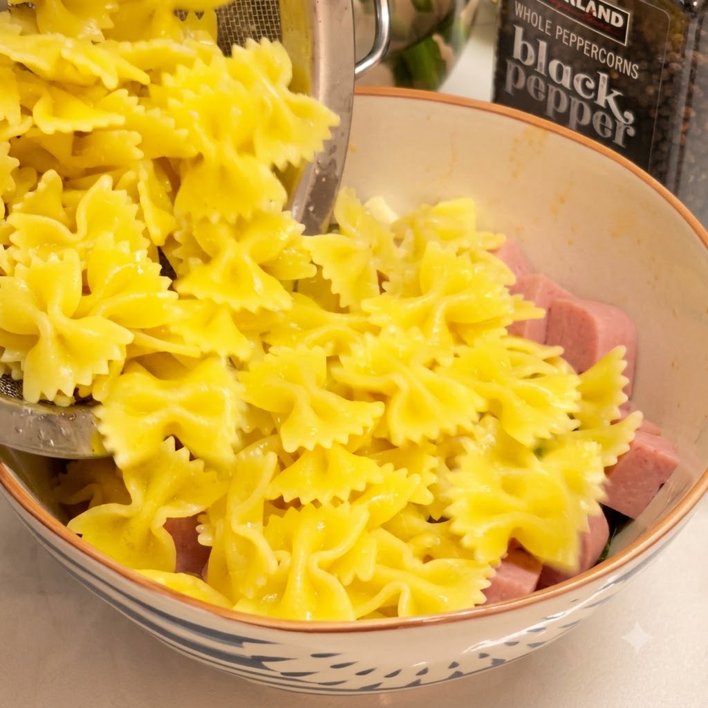 Pasta farfalle cocida siendo vertida desde un colador en un bol grande lleno de jamón en cubos y verduras.