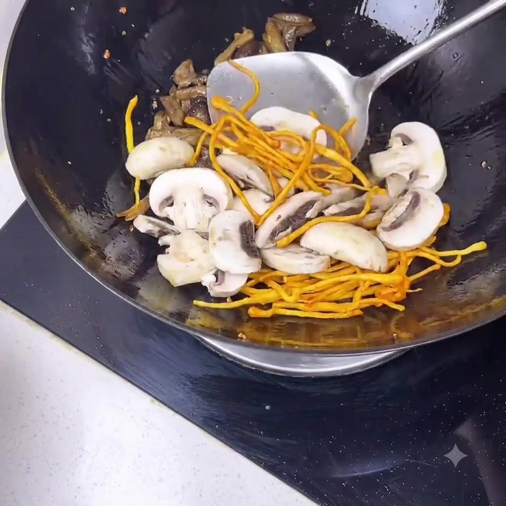 Una espátula de metal removiendo láminas de champiñón blanco y flores de cordyceps amarillas dentro de un wok negro.
