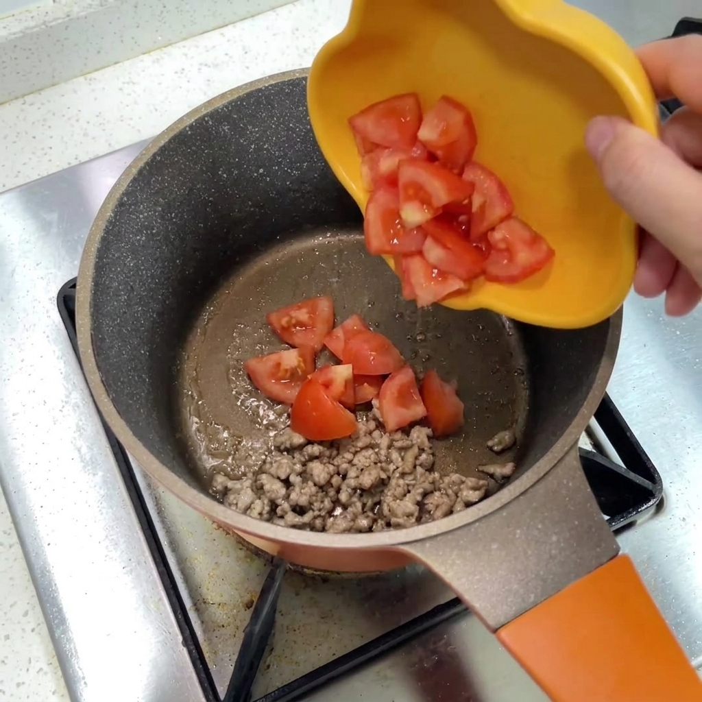 Tomates picados deslizándose desde un plato amarillo hacia una olla con ternera picada.
