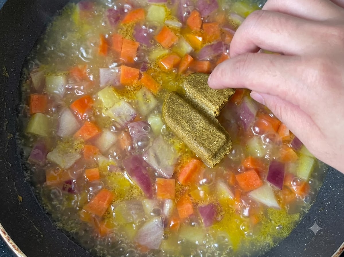 Bloques de curry siendo arrojados a una olla hirviendo de verduras cocidas y caldo.