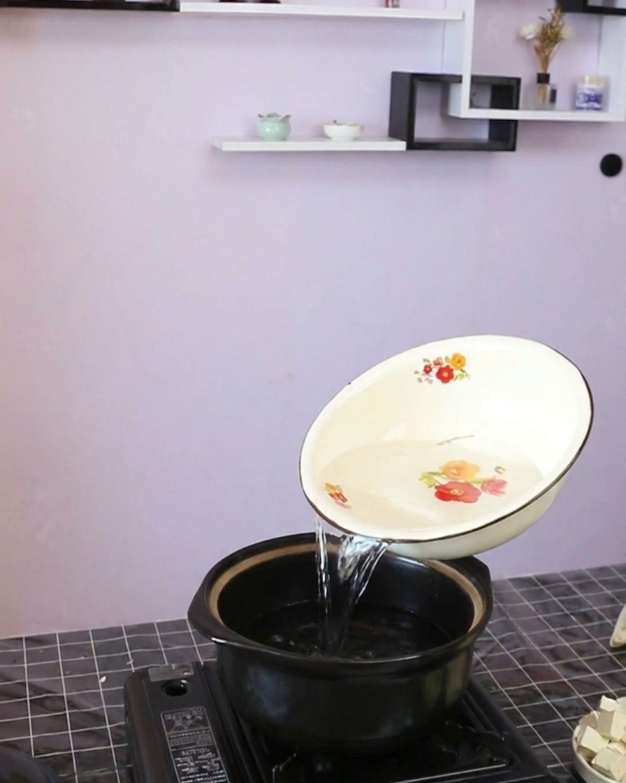 Agua clara siendo vertida desde un cuenco blanco decorado hacia una olla de barro oscura.
