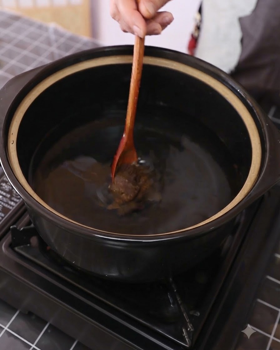Pasta de soja siendo revuelta en agua dentro de una olla oscura usando una cuchara de madera.
