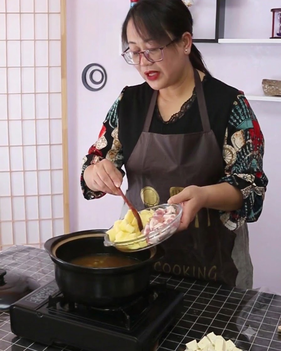 Patatas y panceta de cerdo siendo vertidas desde un plato de vidrio hacia una olla de cerámica negra llena de caldo.