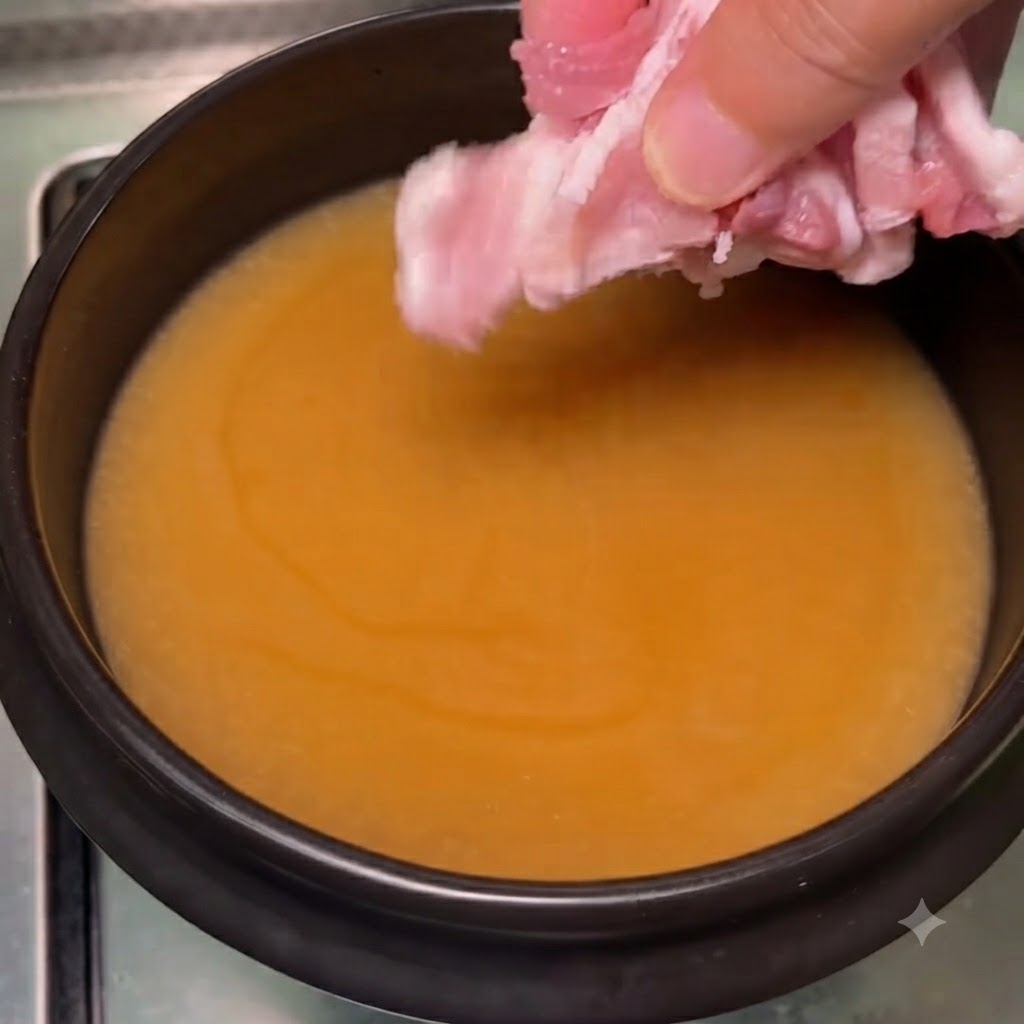 Dedos colocando finas rodajas de panceta de cerdo rosa en el caldo de soja burbujeante.