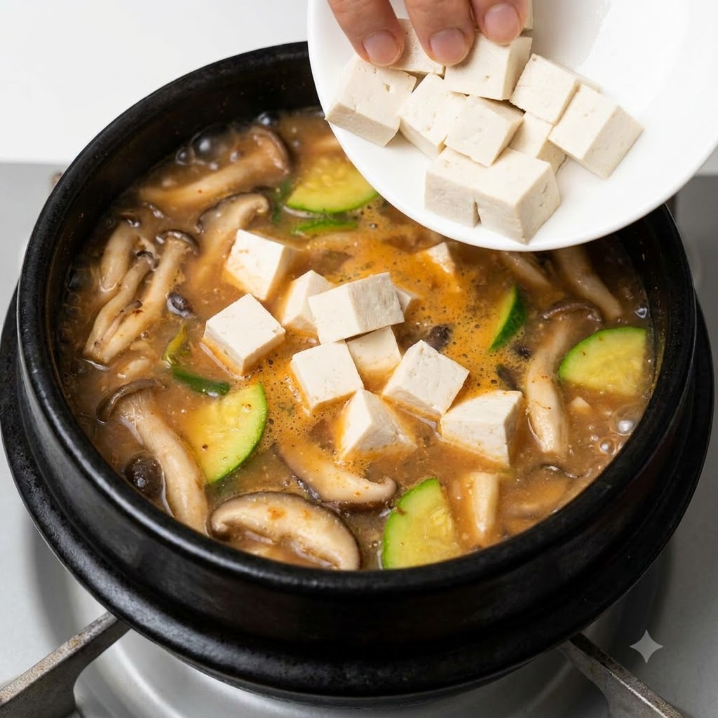 Tofu blanco en cubos siendo añadido desde un pequeño plato blanco en un estofado de pasta de soja coreana burbujeante.