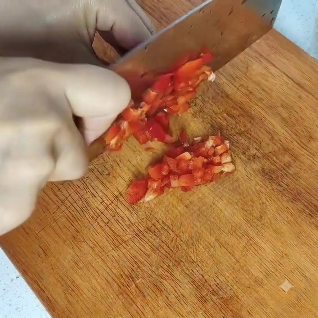 Manos cortando un pimiento rojo en trozos pequeños sobre una tabla de cortar de madera con un cuchillo grande.