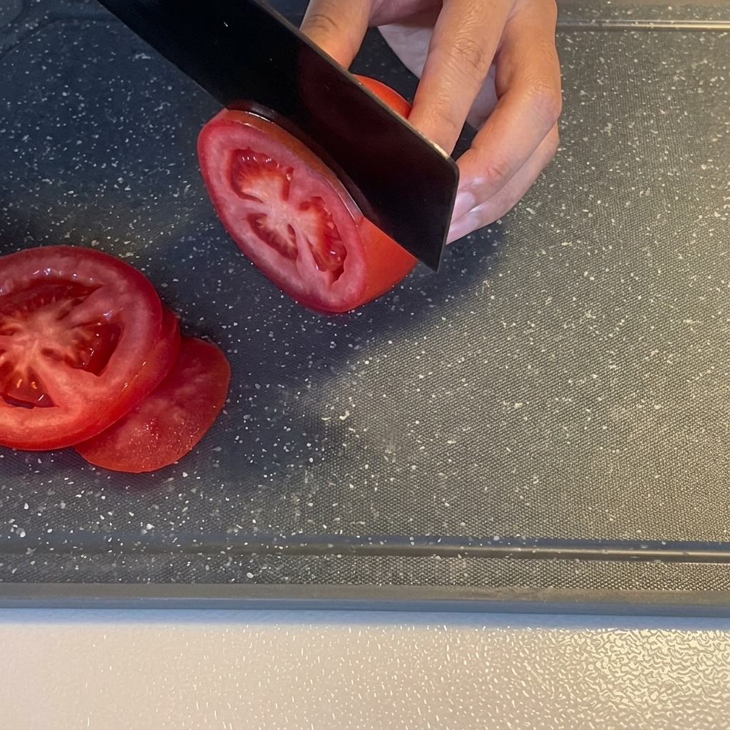 Una mano cortando un tomate rojo fresco en rodajas con un cuchillo negro sobre una tabla de cortar.