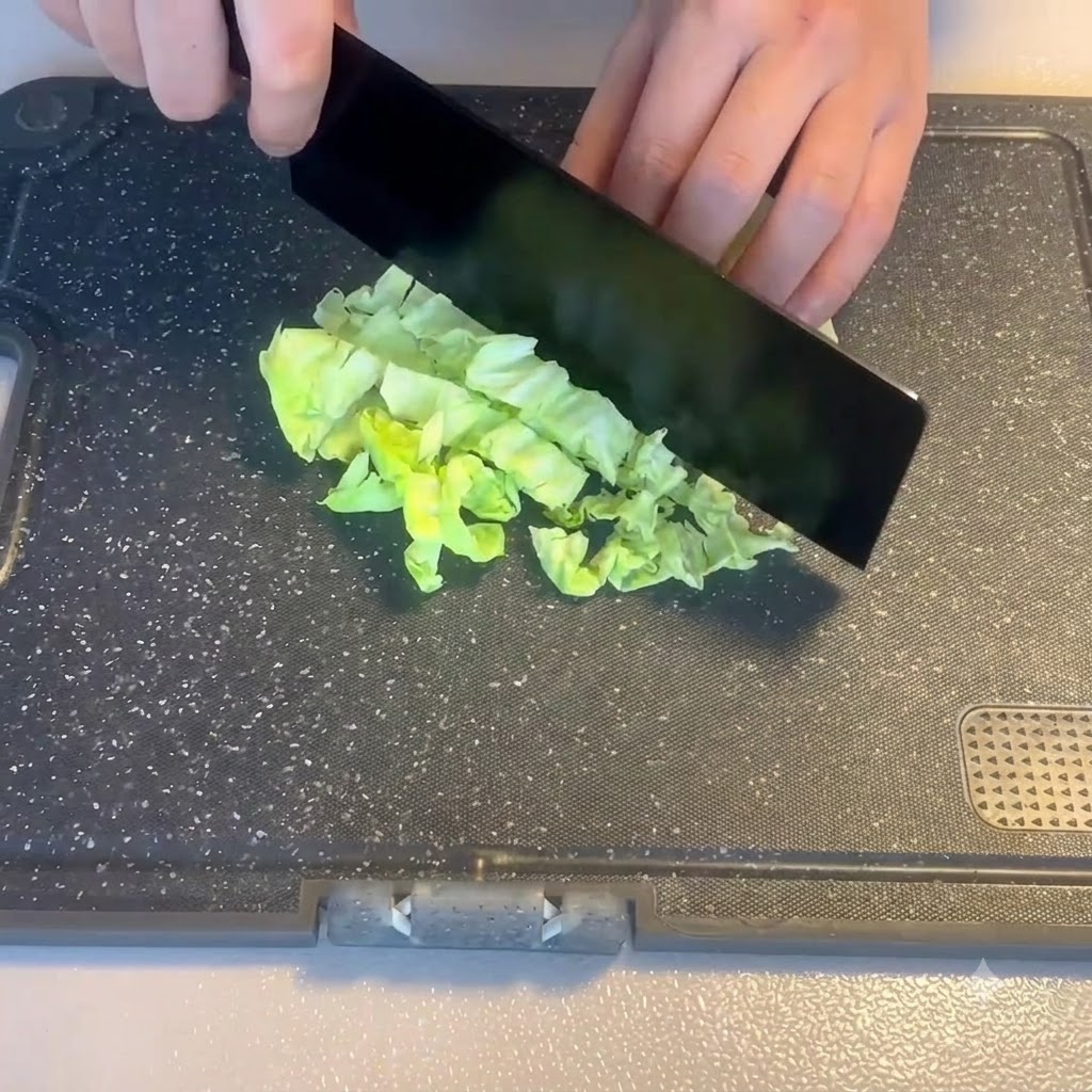 Una mano usando un hachuela negra para picar verduras frescas de hoja verde en una tabla de cortar.