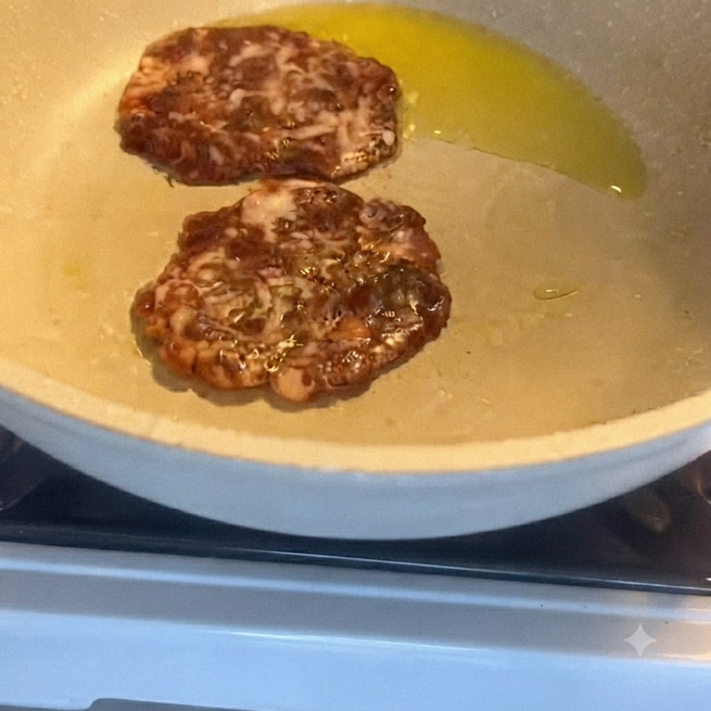 Dos hamburguesas de cerdo cociéndose en una sartén con aceite, con una rodaja de queso colocada sobre una de ellas.