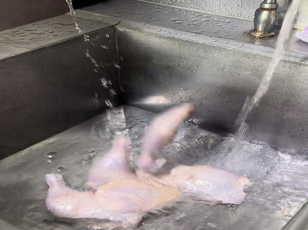 Muslos de pollo enteros crudos lavándose bajo el agua corriente en un fregadero de acero inoxidable.