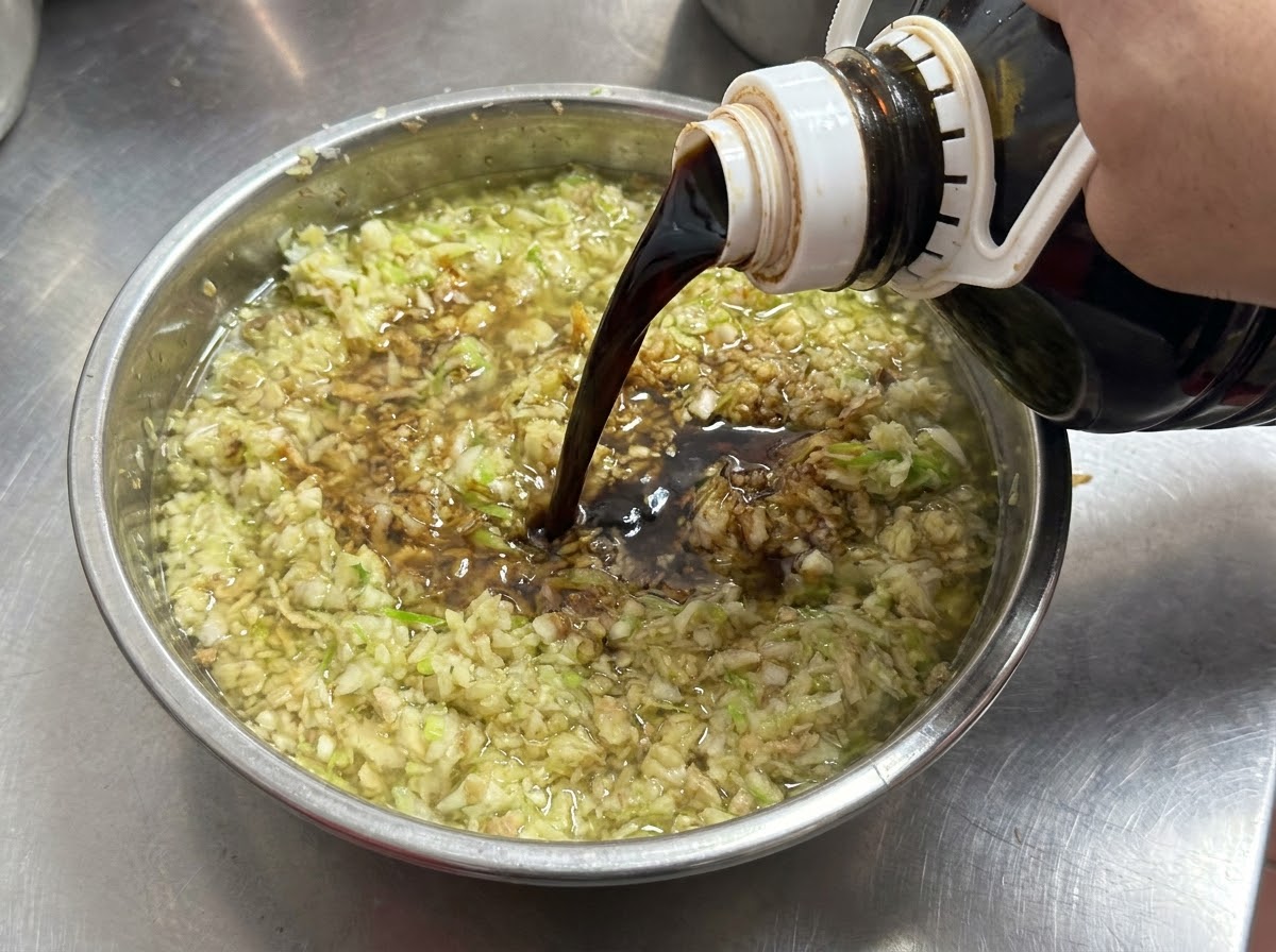 Salsa de soja oscura vertiéndose desde una botella en un bol lleno de ajo picado y cebollinos.