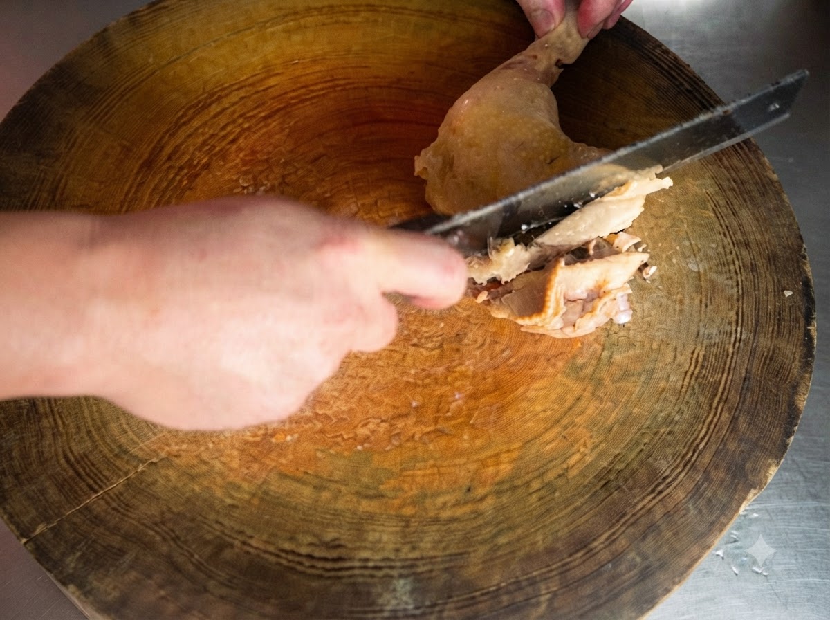 Un cuchillo afilado cortando un muslo de pollo cocido sobre una tabla de cortar de madera redonda.