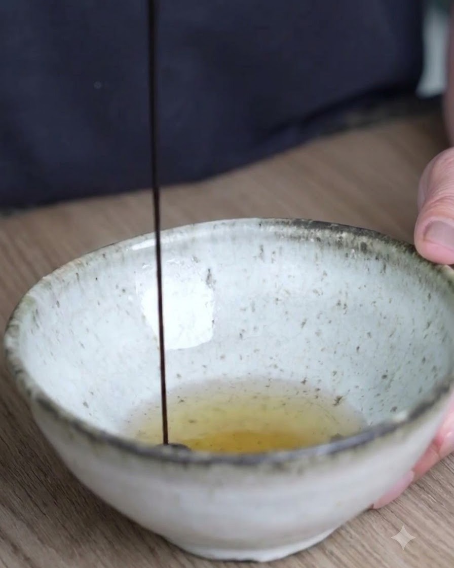 Salsa de soja oscura vertiéndose desde arriba en un tazón de cerámica que contiene el aceite infusionado tibio.