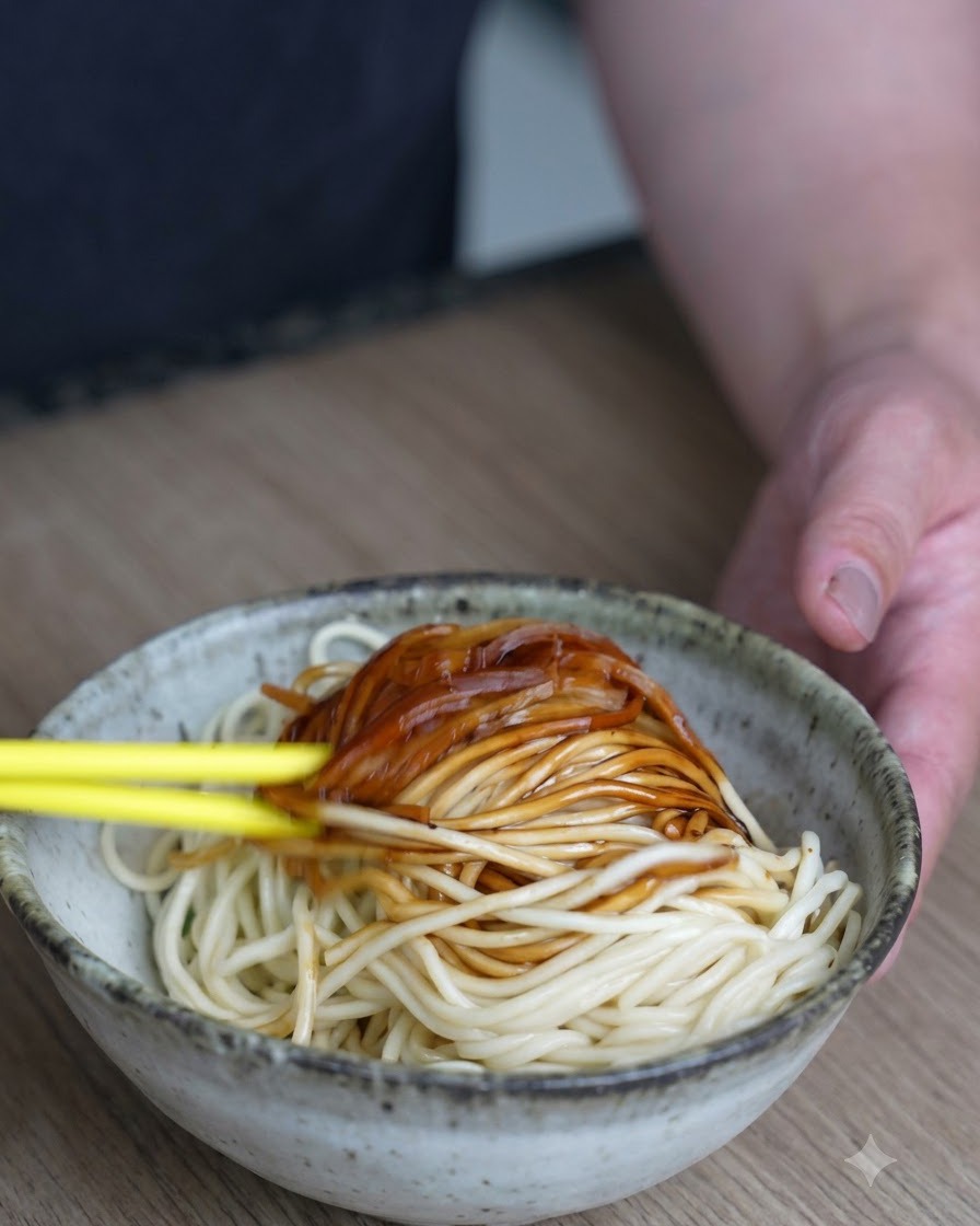 Fideos y salsa oscura siendo revueltos y combinados con palillos amarillos brillantes dentro de un tazón de cerámica.