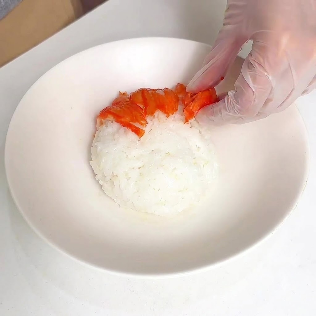 Una mano con guante acomodando palitos de cangrejo cortados en patrón radial sobre un montón de arroz blanco en un tazón.